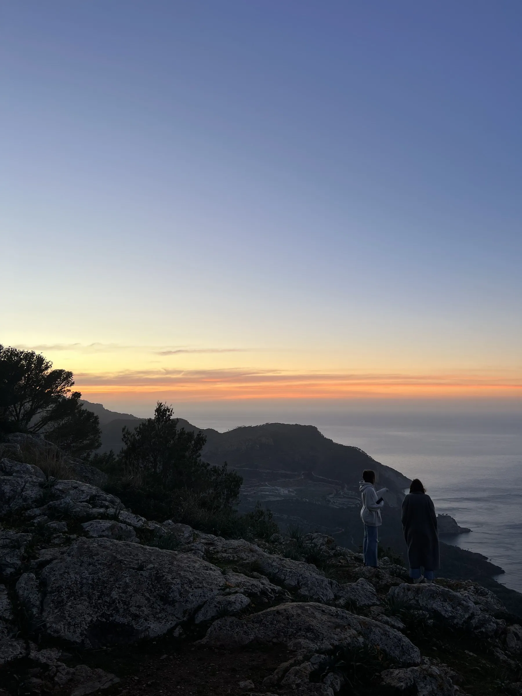 Two people standing on the rocks looking out at the sea off Banyalbufar as the light fades.