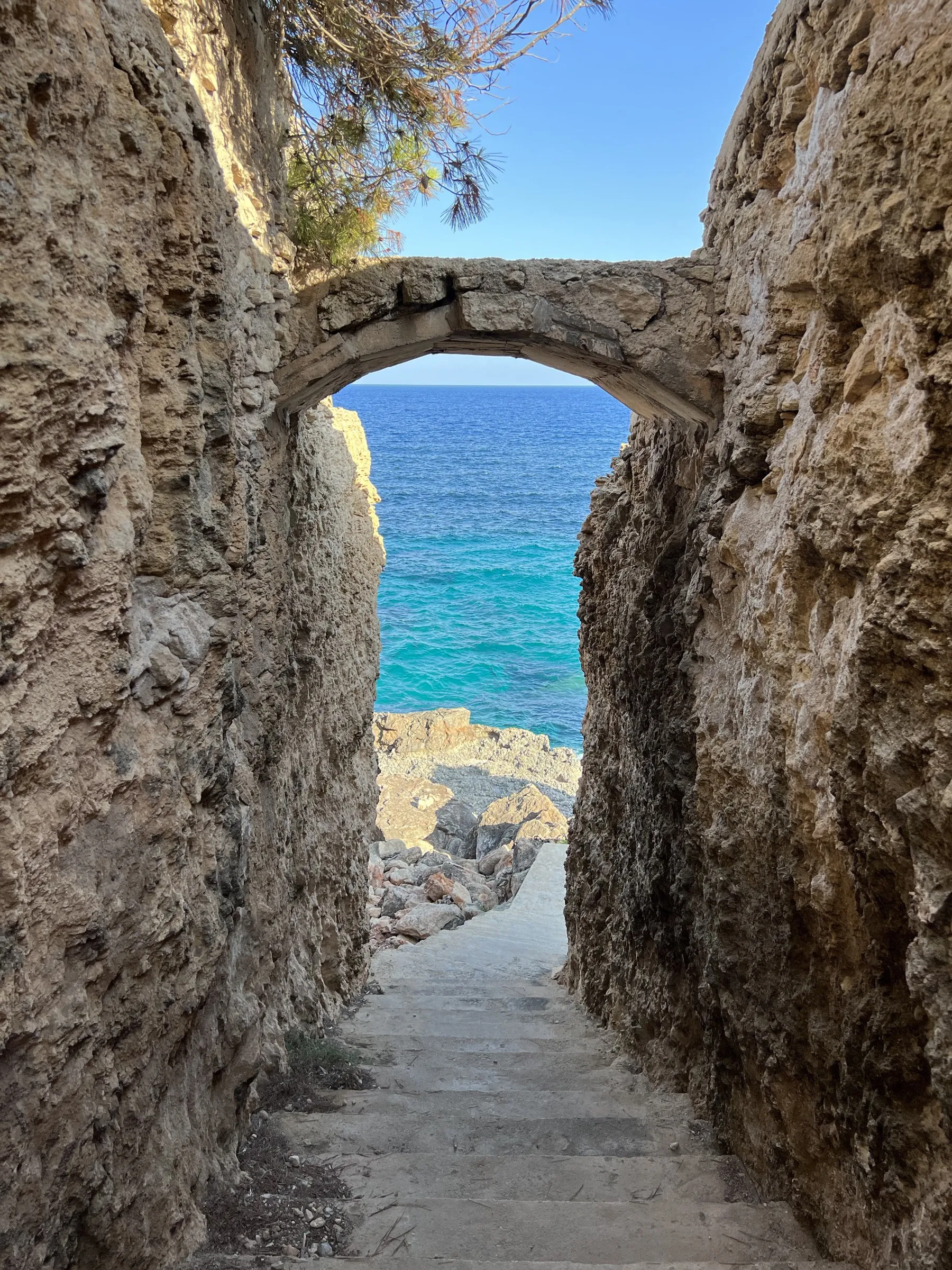 A stone arch cut into the rock, with steps leading down to the turquoise water of a small cove.