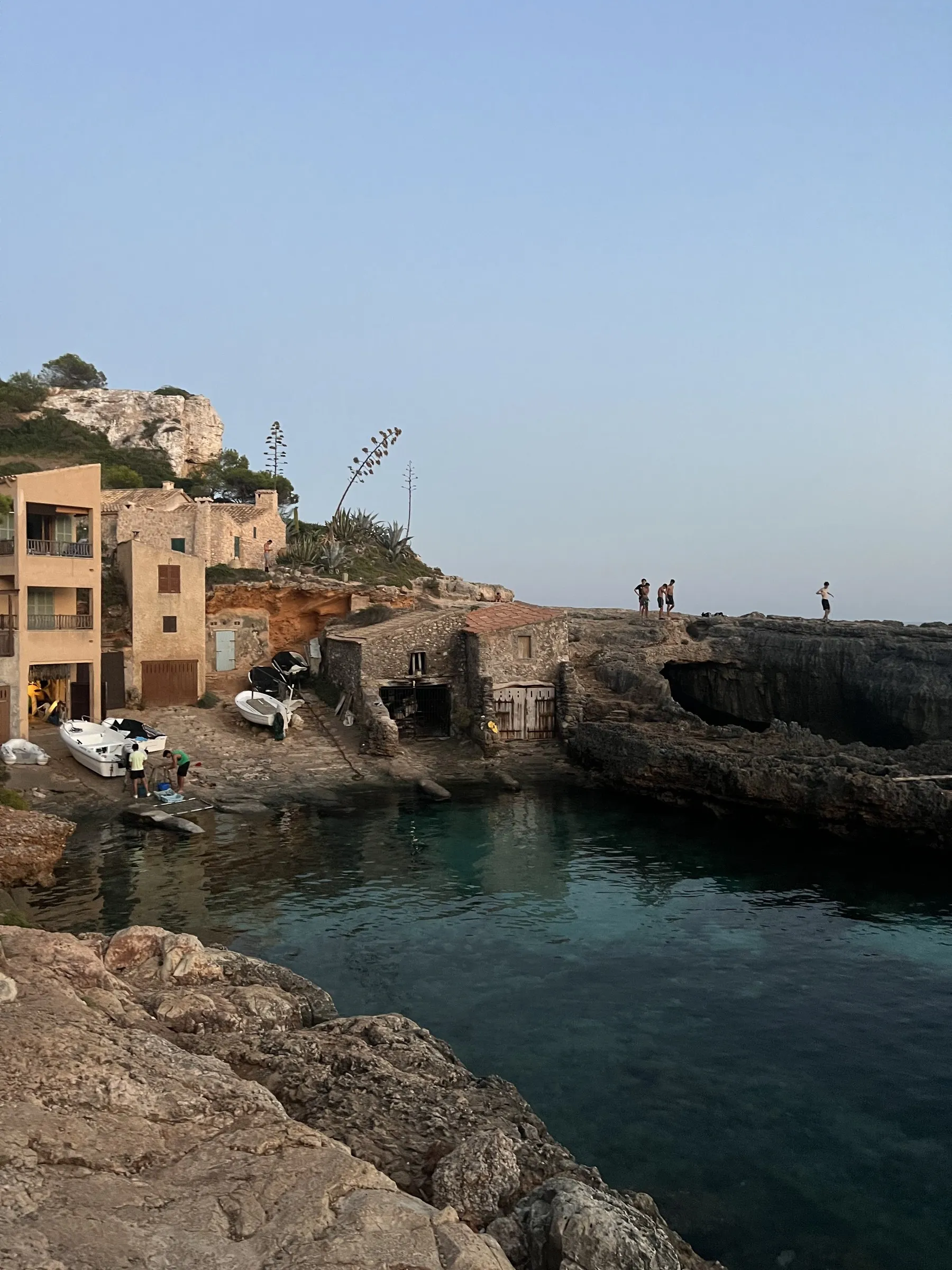 Die steinernen Bootshäuser der Cala s'Almunia am Wasser, mit an Land gezogenen Booten und Menschen auf den Felsen.