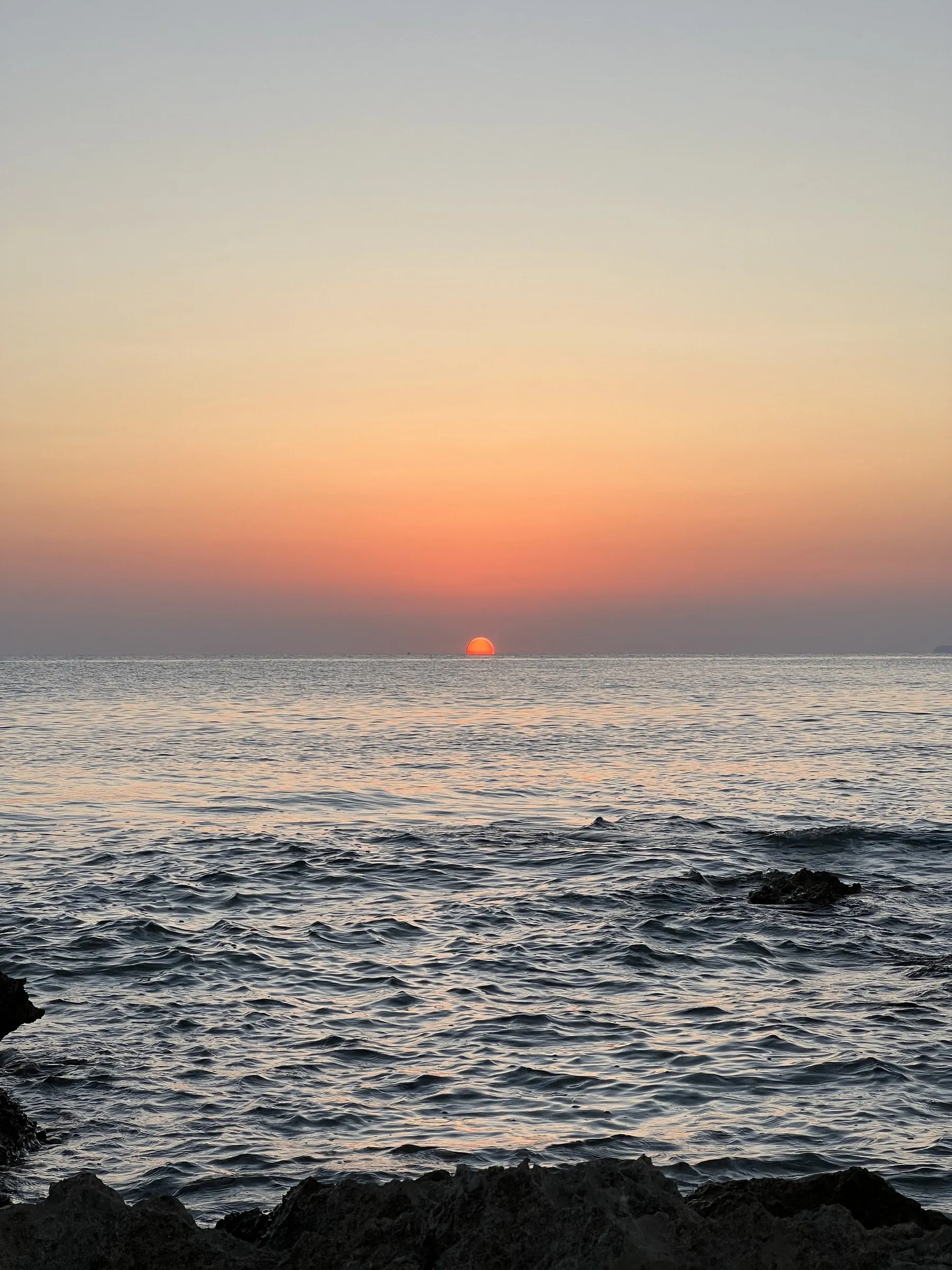 The sun touching the horizon over the open sea off Cap de ses Salines, with low rocks in the foreground.